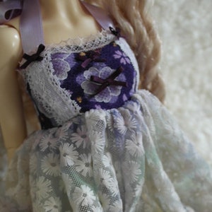 May include: A doll dress featuring a bodice with a purple floral pattern and white lace trim. The skirt is made of a sheer, light blue fabric with a delicate white floral pattern. The dress has lavender ribbon straps.