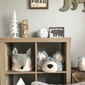 May include: A wooden shelf unit with a white faux fur pillow, a wooden mountain sculpture, a crocheted gray bear head, a crocheted gray fox basket, a wooden sign that says "Baby Bear", a white globe with black stars, a wooden sign that says "Love you to the moon and back", and a wooden bear silhouette.