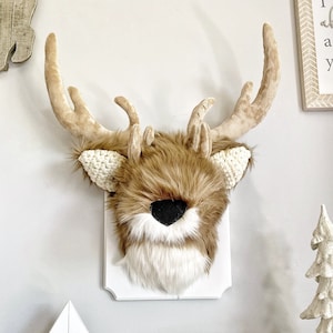 Puede incluir: Una cabeza de ciervo de piel sintética marrón con cuernos blancos montada en una placa de madera blanca. La cabeza de ciervo tiene una nariz negra y piel blanca alrededor de los ojos. La placa está colgada en una pared blanca. Hay un letrero de madera debajo de la cabeza de ciervo que dice "i love you more".