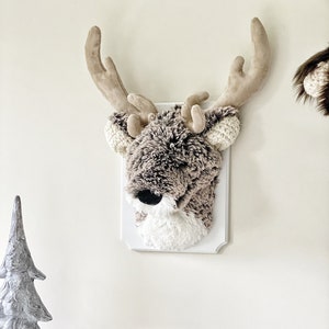 May include: A plush gray and brown reindeer head mounted on a white plaque. The reindeer has large, brown antlers and a white face. The plaque is hanging on a white wall.