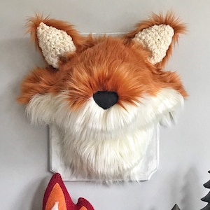 May include: A faux fur fox head wall decoration with orange and white fur, a black nose, and cream-coloured ears. A felt campfire, a hedgehog, and wooden trees decorate a shelf, creating a woodland theme.