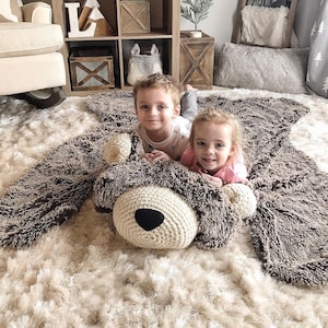May include: A large, plush, bear-shaped rug featuring a cream-colored crocheted snout with a black nose, surrounded by dark brown, shaggy, faux-fur material that forms the head, ears, and body of the bear, laid flat on a light-colored shag carpet.
