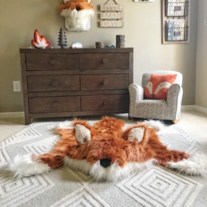 May include: A plush, faux fur rug shaped like a fox, featuring a vibrant orange and white coat with a black nose and textured ears. The rug is laid flat on a light gray carpet with a geometric diamond pattern, showcasing a soft, shaggy texture.