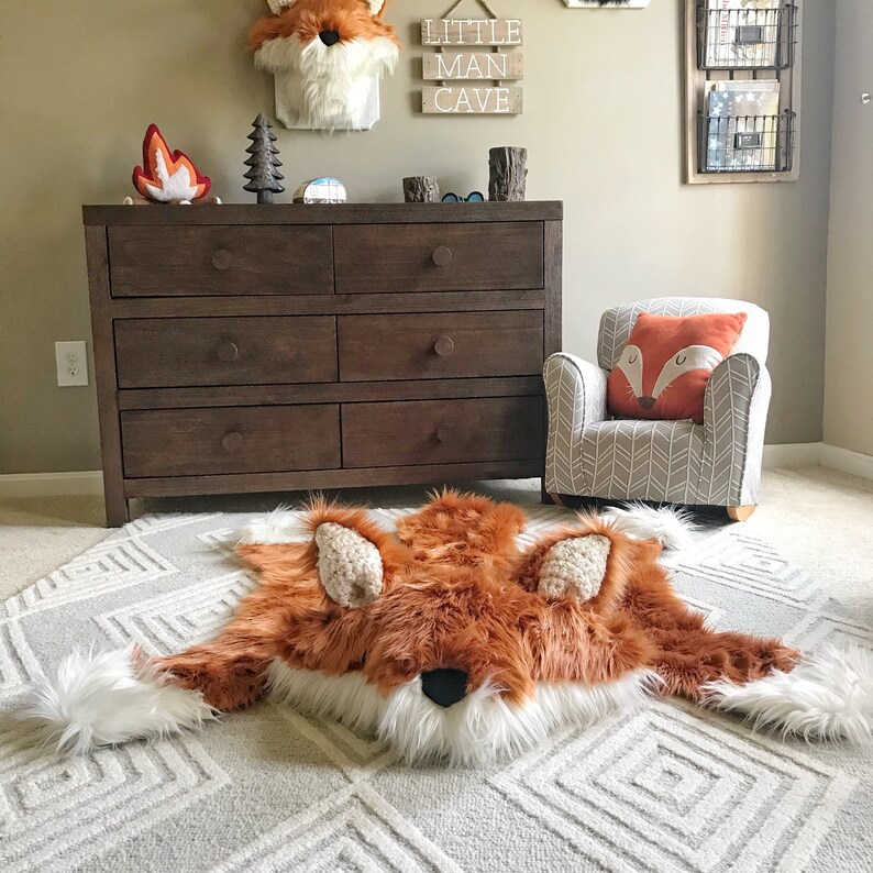 May include: A plush, faux fur rug shaped like a fox, featuring a vibrant orange and white coat with a black nose and textured ears. The rug is laid flat on a light gray carpet with a geometric diamond pattern, showcasing a soft, shaggy texture.
