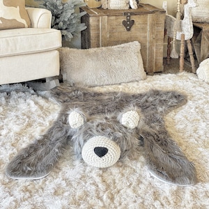 May include: A faux fur area rug shaped like a bear, featuring long grey synthetic fur, a cream-colored crocheted snout with a black triangular nose, and two cream-colored crocheted ears. The rug lies flat with extended limbs.