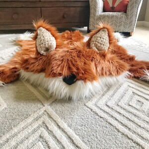 May include: A plush fox-shaped rug featuring long, shaggy rust-orange faux fur, a white faux fur chest and muzzle, and two textured cream-colored crocheted ears. The fox has a small, solid black nose centered on the white fur section.
