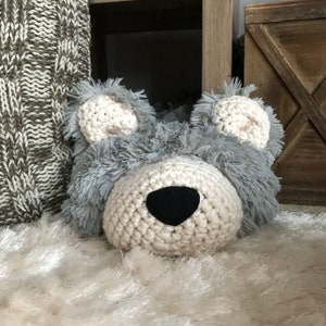 May include: A crocheted gray and white stuffed animal shaped like a bear's head. The bear has a black nose and is sitting on a white fluffy rug.