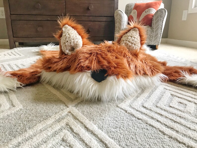 May include: A plush fox-shaped rug featuring long, rust-orange faux fur, a white faux fur chest and muzzle area, two upright crocheted ears with textured beige centers, and a small, triangular black nose centered on the face.