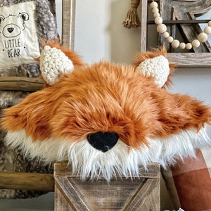 May include: A plush fox head pillow with a white fur trim and black nose. The pillow is sitting on a wooden crate.