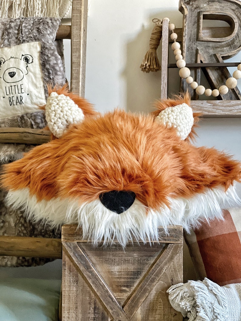 May include: A plush fox head pillow with a white fur trim and black nose. The pillow is sitting on a wooden crate.