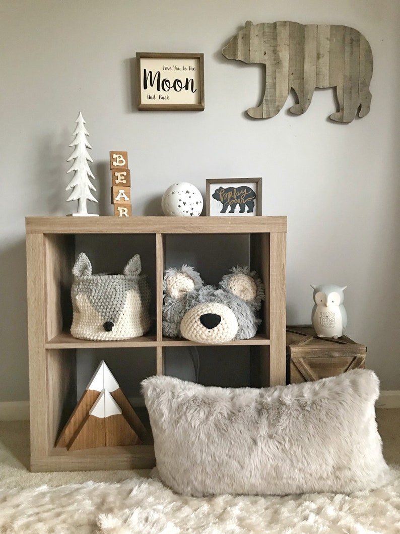 May include: A wooden shelf unit with a white faux fur pillow, a wooden mountain sculpture, a crocheted gray bear head, a crocheted gray fox basket, a wooden sign that says "Baby Bear", a white globe with black stars, a wooden sign that says "Love you to the moon and back", and a wooden bear silhouette.