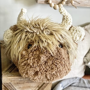 Puede incluir: Una cabeza de vaca de las tierras altas de peluche de crochet marrón y blanco con una textura de pelo marrón esponjosa. La cabeza de vaca está sentada en un estante de madera con una manta blanca y gris en el fondo.