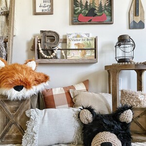 May include: A nursery room with a wooden shelf holding a book titled "A Visitor for Bear". There are two pillows on the floor, one with a fox face and the other with a bear face. The wall has a wooden sign with the text "Nap Build Destroy Repeat" and a framed picture of a bear in a canoe.