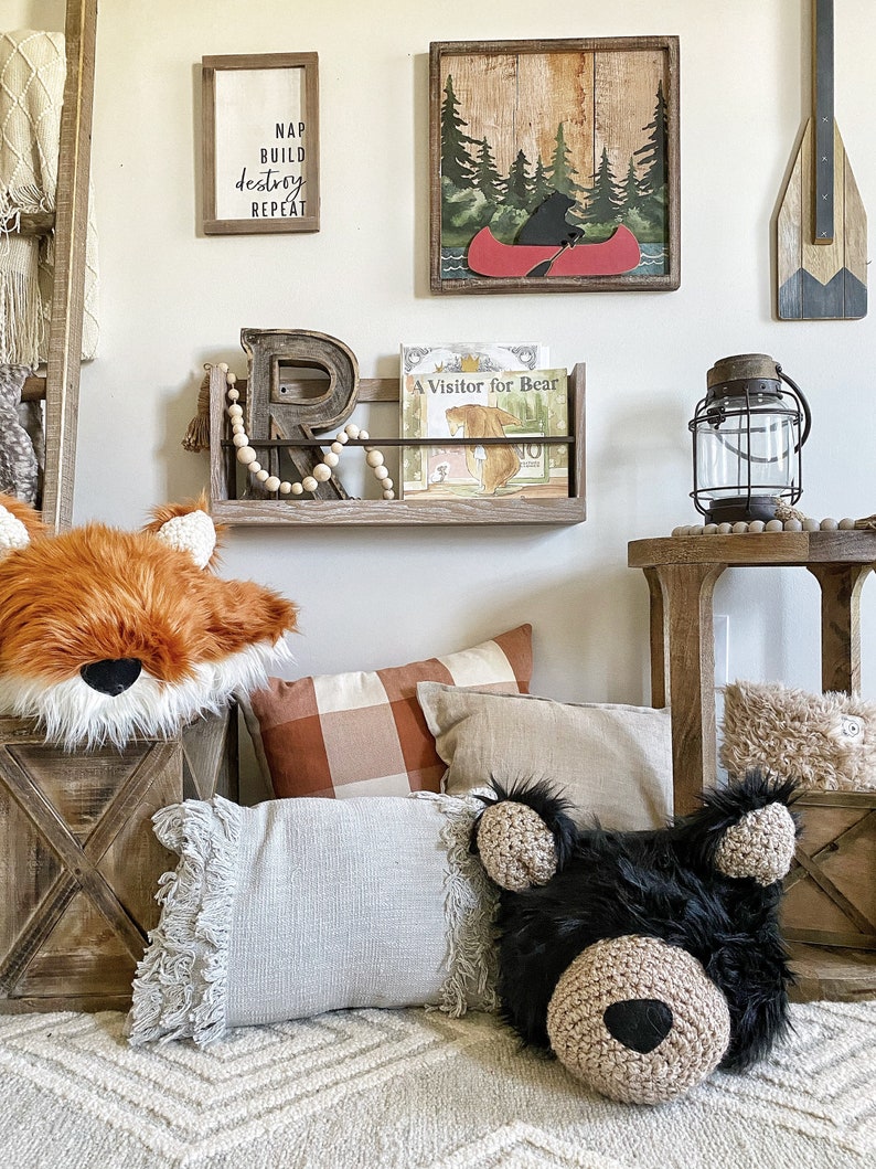 May include: A nursery room with a wooden shelf holding a book titled "A Visitor for Bear". There are two pillows on the floor, one with a fox face and the other with a bear face. The wall has a wooden sign with the text "Nap Build Destroy Repeat" and a framed picture of a bear in a canoe.