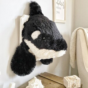 Op de afbeelding: Een zwart-witte pluche orka-kop gemonteerd aan een witte muur met een houten plank eronder. De plank heeft een witte schelp en een klein houten doosje met een touw- en wielontwerp. Een ingelijst print met de tekst "I absolutely adore you" hangt aan de muur achter de orka.