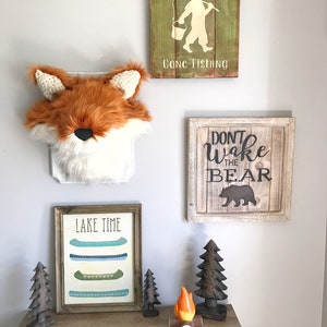 May include: A rustic wall decor arrangement featuring a faux fox head, framed signs, and wooden accents. The signs read "Gone Fishing" and "Don't Wake the Bear." A framed print displays canoes with the text "Lake Time."