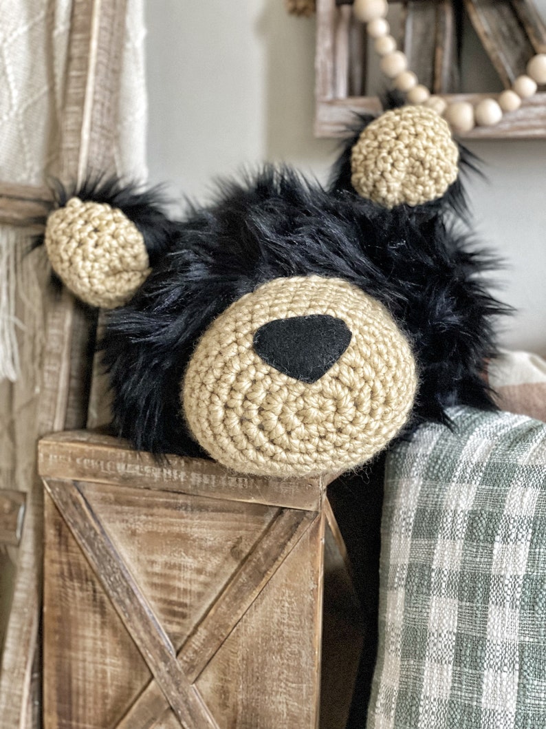 May include: A crocheted black bear head with a tan crocheted nose and ears. The bear head is sitting on a wooden crate with a green and white plaid blanket behind it.