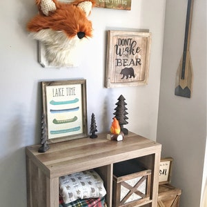 May include: A rustic-themed room decor featuring a fox head wall hanging, wooden signs with text, and a wooden storage unit. The signs read "Gone Fishing" and "Don't Wake the Bear." The unit displays blankets, a bear-shaped basket, and decorative wooden trees.