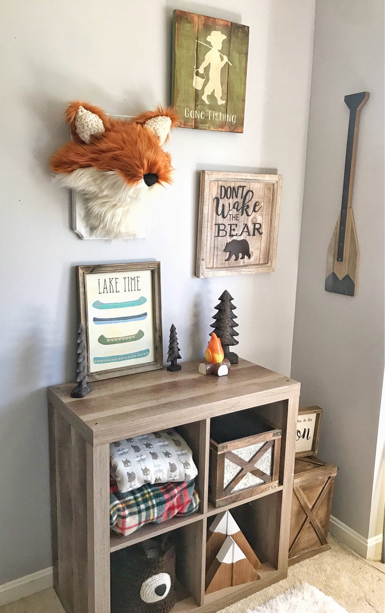 May include: A rustic-themed room decor featuring a fox head wall hanging, wooden signs with text, and a wooden storage unit. The signs read "Gone Fishing" and "Don't Wake the Bear." The unit displays blankets, a bear-shaped basket, and decorative wooden trees.