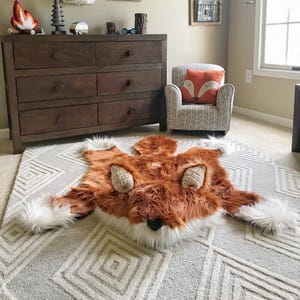 May include: A plush fox-shaped rug featuring long, soft orange-brown faux fur, white accents on the paws and face, and textured beige ears. The rug is laid flat on a light gray area rug with a white geometric diamond pattern across the surface.