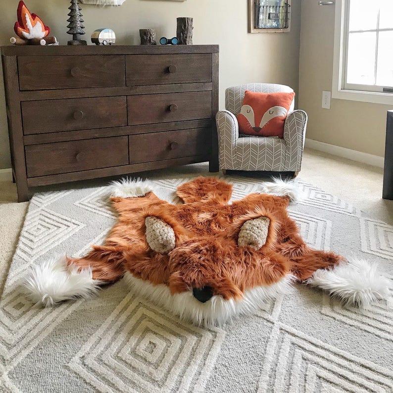 May include: A plush fox-shaped rug featuring long, soft orange-brown faux fur, white accents on the paws and face, and textured beige ears. The rug is laid flat on a light gray area rug with a white geometric diamond pattern across the surface.