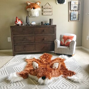 May include: A plush, faux fur fox-shaped rug featuring a vibrant orange and white color palette with textured ears, resting on a light gray geometric patterned carpet in a room with a dark brown wooden dresser and a small upholstered armchair.