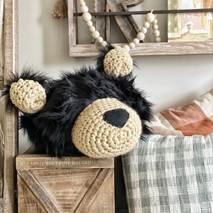 May include: A crocheted black bear head with brown yarn details and black eyes. The bear head is sitting on a wooden box with a cross pattern. The box is in front of a plaid blanket and a wooden shelf with a wooden bead garland.