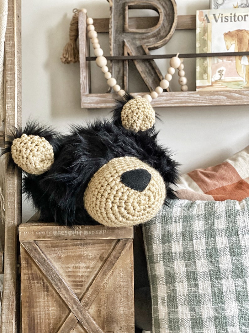 May include: A crocheted black bear head with brown yarn details and black eyes. The bear head is sitting on a wooden box with a cross pattern. The box is in front of a plaid blanket and a wooden shelf with a wooden bead garland.