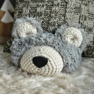 May include: A crocheted gray and white bear head with a black nose. The bear head is sitting on a white fluffy surface.