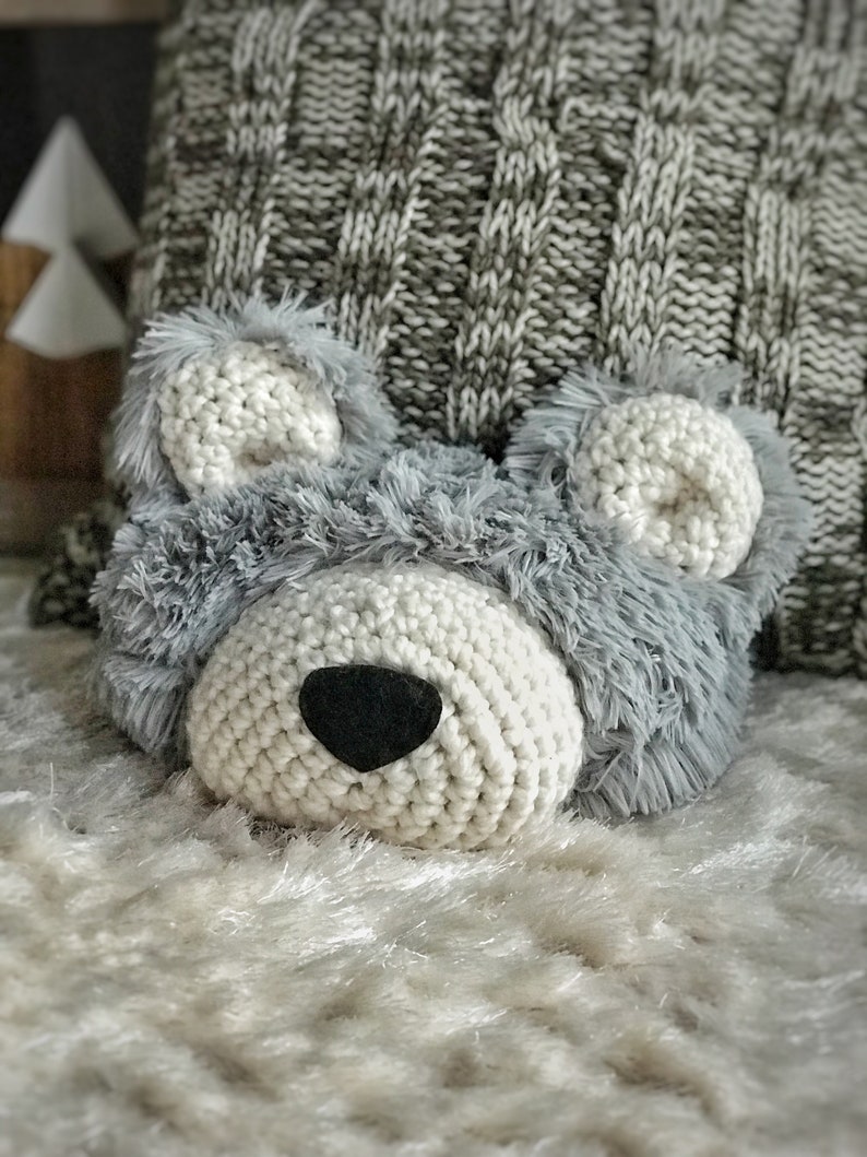 May include: A crocheted gray and white bear head with a black nose. The bear head is sitting on a white fluffy surface.