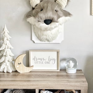 May include: A faux fur wolf head wall decor with a white plaque. A wooden sign with the text "dream big LITTLE ONE" sits on a wooden table, along with a wooden moon and a snow globe. The decor has a neutral color palette.