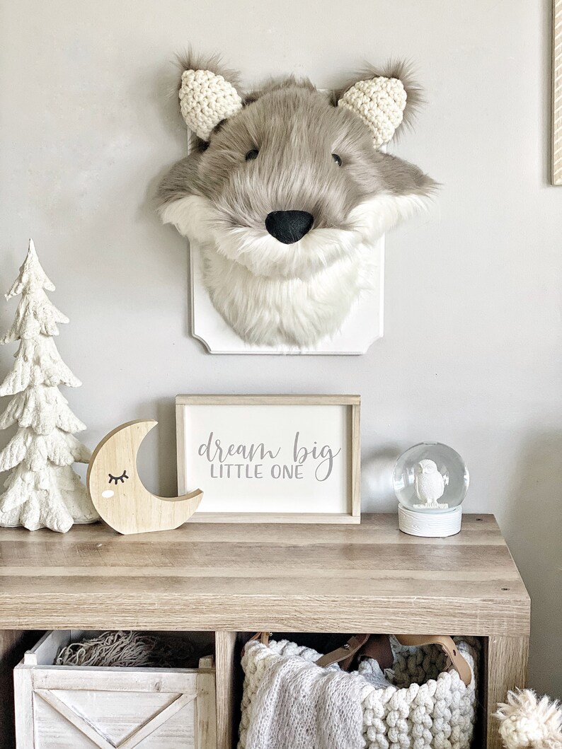 May include: A faux fur wolf head wall decor with a white plaque. A wooden sign with the text "dream big LITTLE ONE" sits on a wooden table, along with a wooden moon and a snow globe. The decor has a neutral color palette.