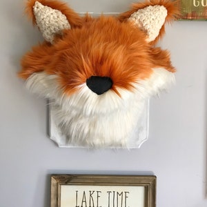 May include: A faux fox head wall decor with orange, white, and black fur. The fox head has crocheted ears and is mounted on a clear, decorative backing. The wall art is displayed on a light gray wall.