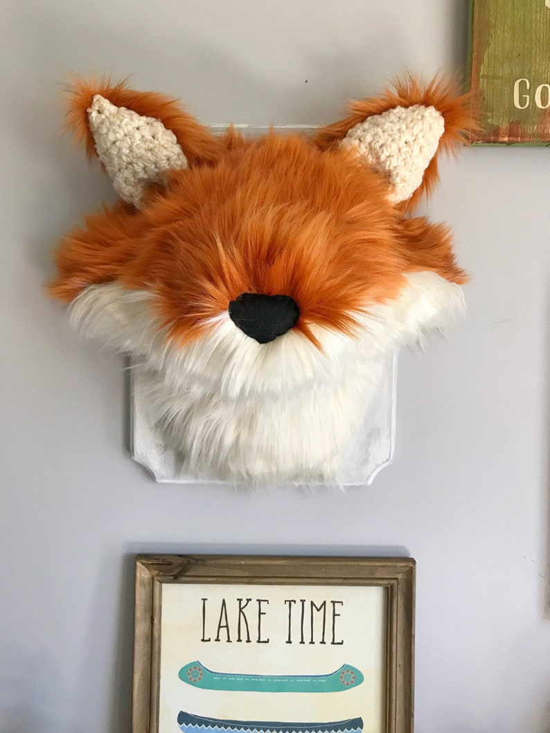 May include: A faux fox head wall decor with orange, white, and black fur. The fox head has crocheted ears and is mounted on a clear, decorative backing. The wall art is displayed on a light gray wall.