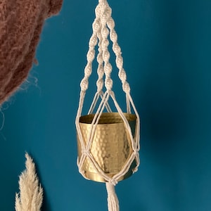 May include: A white macrame plant hanger with a gold metal pot. The hanger has a tassel at the bottom.