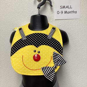 May include: A yellow and black bee-themed baby bib with a black and white bow tie. The bib has a red button nose and black eyes. The bib is labeled "SMALL 0-9 Months".