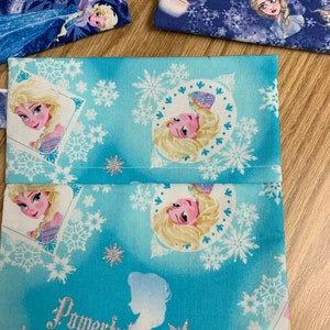 Op de afbeelding: Een blauwe stoffen tas met een wit sneeuwvlokkenpatroon en afbeeldingen van Elsa uit de film Frozen. De tas heeft een naad in het midden en de woorden "Powerful" en "ID" staan ​​onderaan gedrukt.