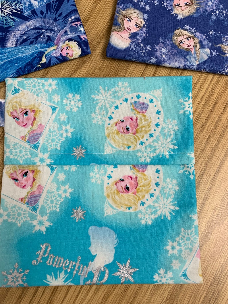 Op de afbeelding: Een blauwe stoffen tas met een wit sneeuwvlokkenpatroon en afbeeldingen van Elsa uit de film Frozen. De tas heeft een naad in het midden en de woorden "Powerful" en "ID" staan ​​onderaan gedrukt.