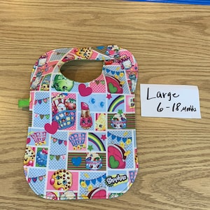 May include: A colorful Shopkins-themed bib with a green trim and a neck opening. The bib has a patchwork design with various Shopkins characters and objects, including a shopping cart, a rainbow, and a heart. The bib is labeled "Large 6-18 Months".