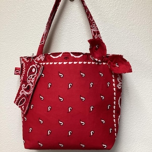 May include: Red bandana tote bag with white paisley pattern and matching handles. The bag has a small, decorative bow on the side and a small bandana attached to the side. The bag is hanging on a silver hook.