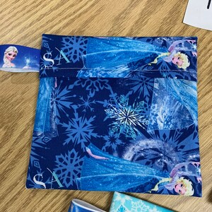 Op de afbeelding: Een blauwe stoffen tas met een ritssluiting en een sneeuwvlokkenpatroon. De tas heeft een afbeelding van Elsa uit de film Frozen. De tas heeft een blauw lint met een klein beeld van Elsa erop.