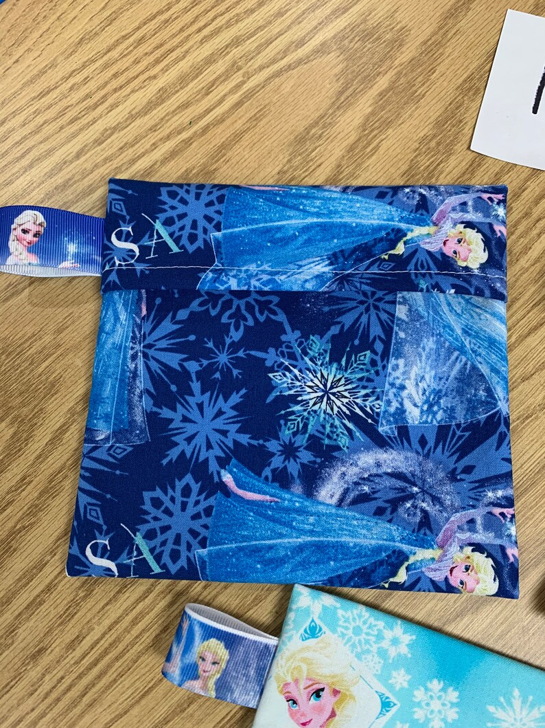 Op de afbeelding: Een blauwe stoffen tas met een ritssluiting en een sneeuwvlokkenpatroon. De tas heeft een afbeelding van Elsa uit de film Frozen. De tas heeft een blauw lint met een klein beeld van Elsa erop.