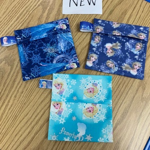Op de afbeelding: Drie kleine stoffen tasjes met een Frozen-thema. De tasjes zijn blauw en lichtblauw met afbeeldingen van Elsa en Anna. De tasjes hebben een ritssluiting en een stoffen lus om op te hangen.