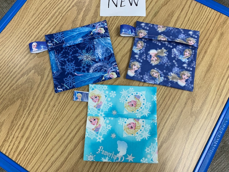 Op de afbeelding: Drie kleine stoffen tasjes met een Frozen-thema. De tasjes zijn blauw en lichtblauw met afbeeldingen van Elsa en Anna. De tasjes hebben een ritssluiting en een stoffen lus om op te hangen.
