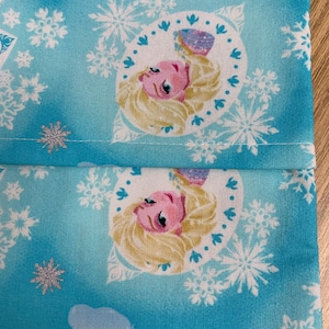 Op de afbeelding: Blauwe stof met een wit sneeuwvlokkenpatroon en een herhaald beeld van een blond meisje met een roze jurk en blauwe ogen. Het meisje bevindt zich in een cirkel met een witte achtergrond.