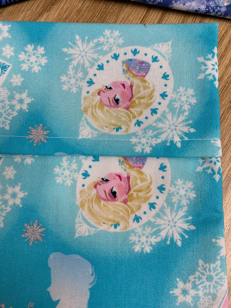 Op de afbeelding: Blauwe stof met een wit sneeuwvlokkenpatroon en een herhaald beeld van een blond meisje met een roze jurk en blauwe ogen. Het meisje bevindt zich in een cirkel met een witte achtergrond.