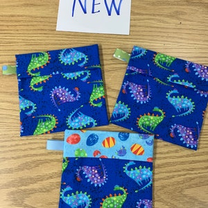 May include: Three fabric pouches with a blue dinosaur pattern and a light blue tab. One pouch has a light blue and egg pattern. The word "NEW" is written in blue marker on a white piece of paper.