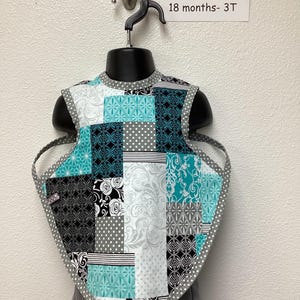 May include: A turquoise, black, white, and gray patchwork bib with a white polka dot trim. The bib is on a hanger and has a label that reads "EXTRA-LARGE 18 months - 3T".