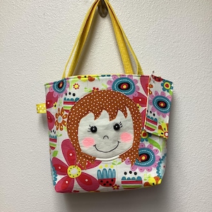 May include: A colorful tote bag with a cheerful girl face applique. The bag features a floral pattern in pink, blue, and yellow, with yellow handles. A small pocket is visible on the side. The bag is hanging on a wall.