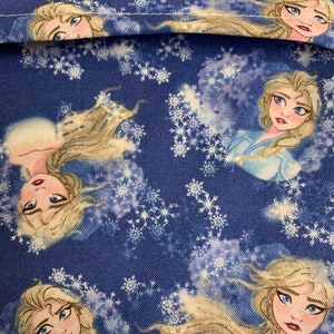 Op de afbeelding: Blauwe stof met een herhalend patroon van een cartoonfiguur met lang blond haar en een blauwe jurk. De achtergrond is een blauw en wit sneeuwvlokkenpatroon.